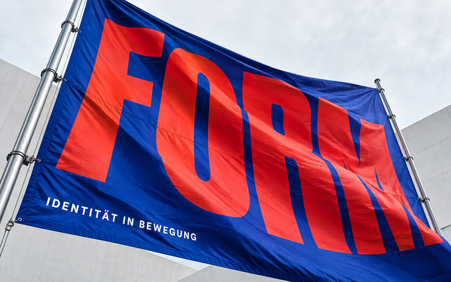 Kobaltblaues Stoffbanner mit roter Markentypografie, im Wind vor einem weißen Industriegebäude